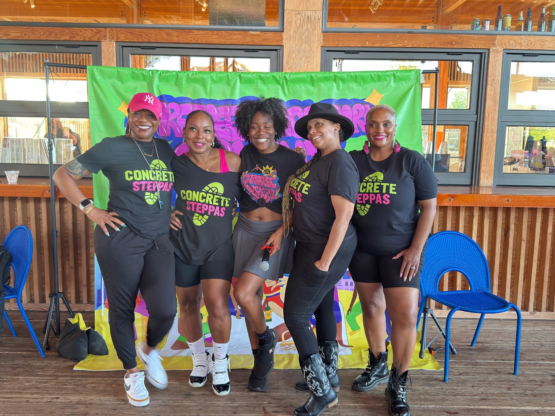 Five people pose together indoors, wearing black Concrete Steppas shirts with pink and green lettering.