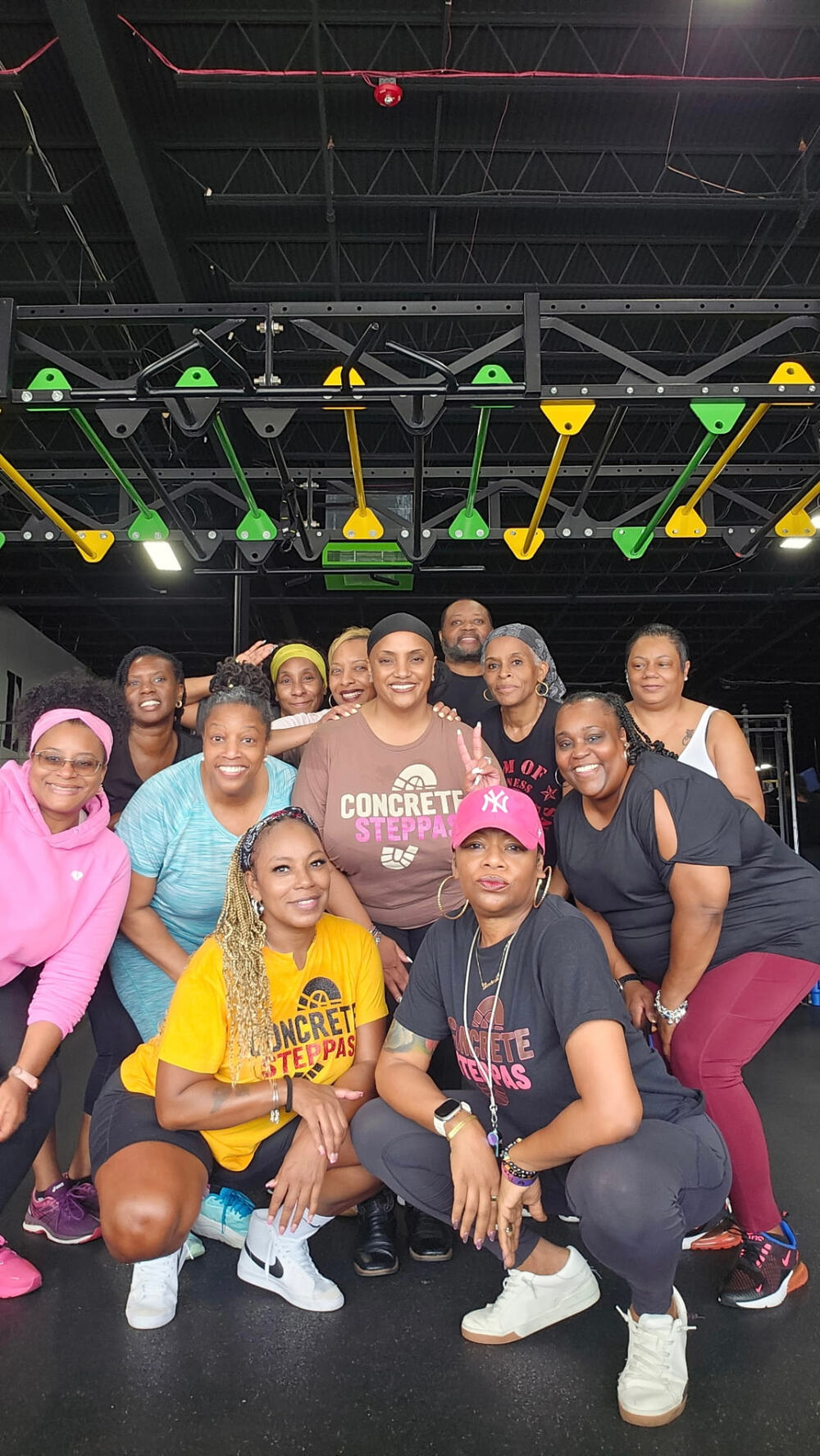 A group poses together in a gym. Several people wear Concrete Steppas™ shirts, with others in colorful workout clothes.